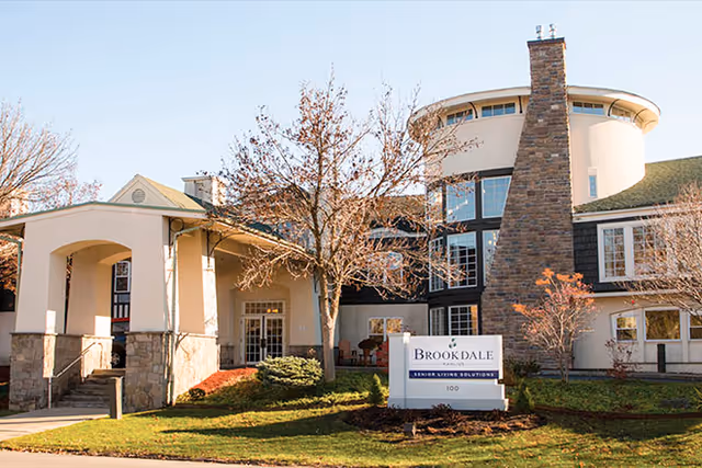 Front exterior of the Brookdale senior living building showing the main entrance, chimney tower, landscaping, and a Brookdale sign.