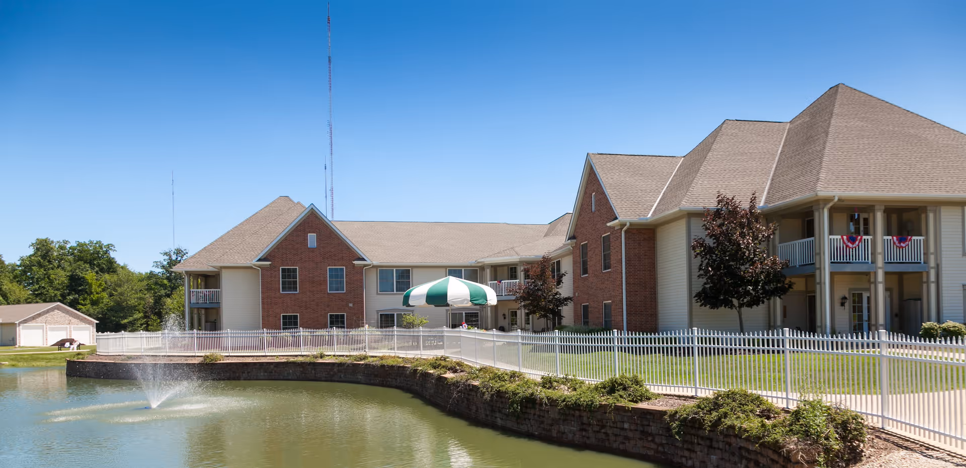 Brick-and-siding senior living building with balconies and a green-and-white awning overlooking a pond with a fountain under a clear blue sky.