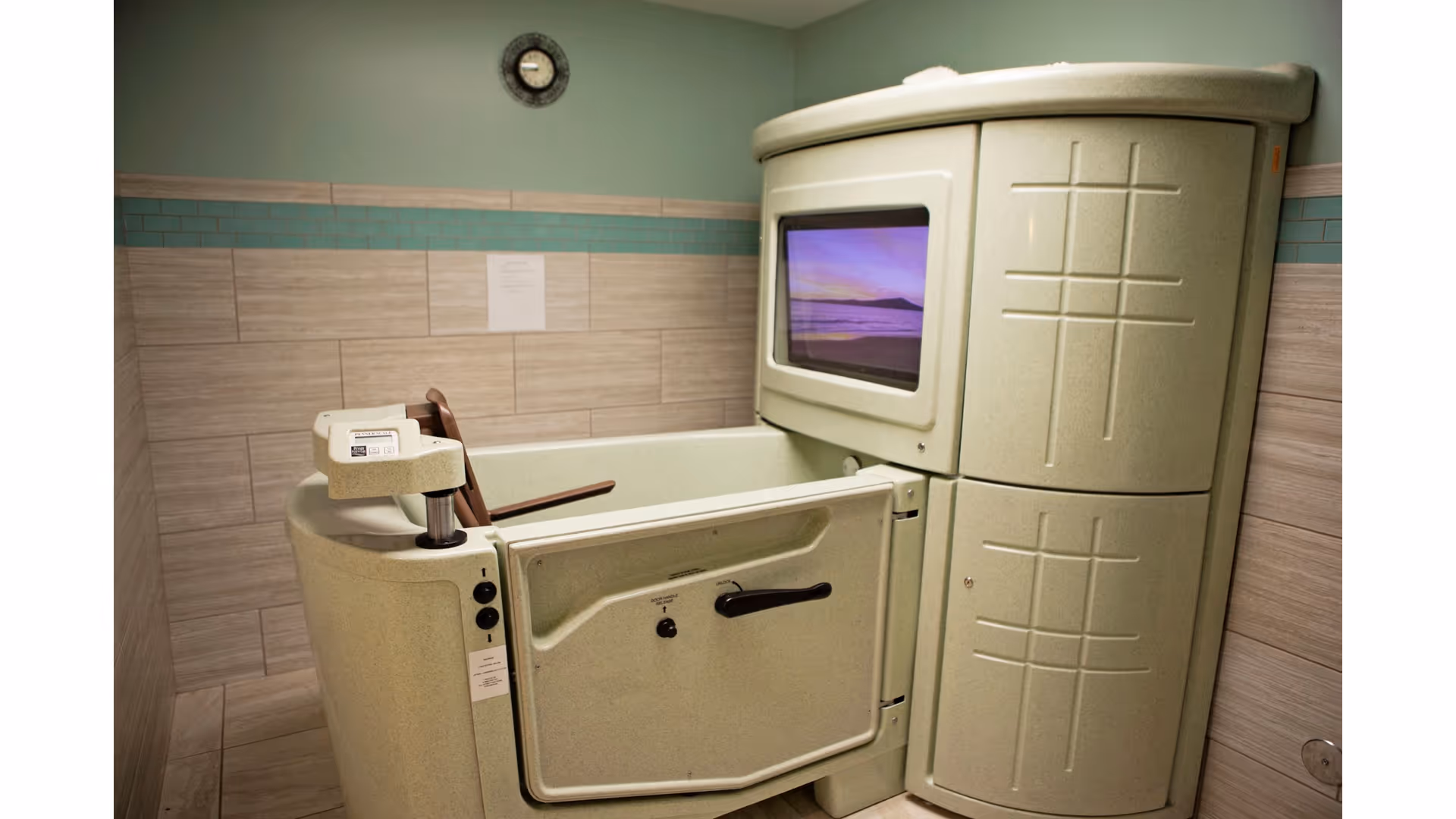 A specialized bathing tub designed for assisted bathing in a senior living facility bathroom, featuring a door for easy access and a built-in screen displaying a scenic image, with tiled walls and a clock above.