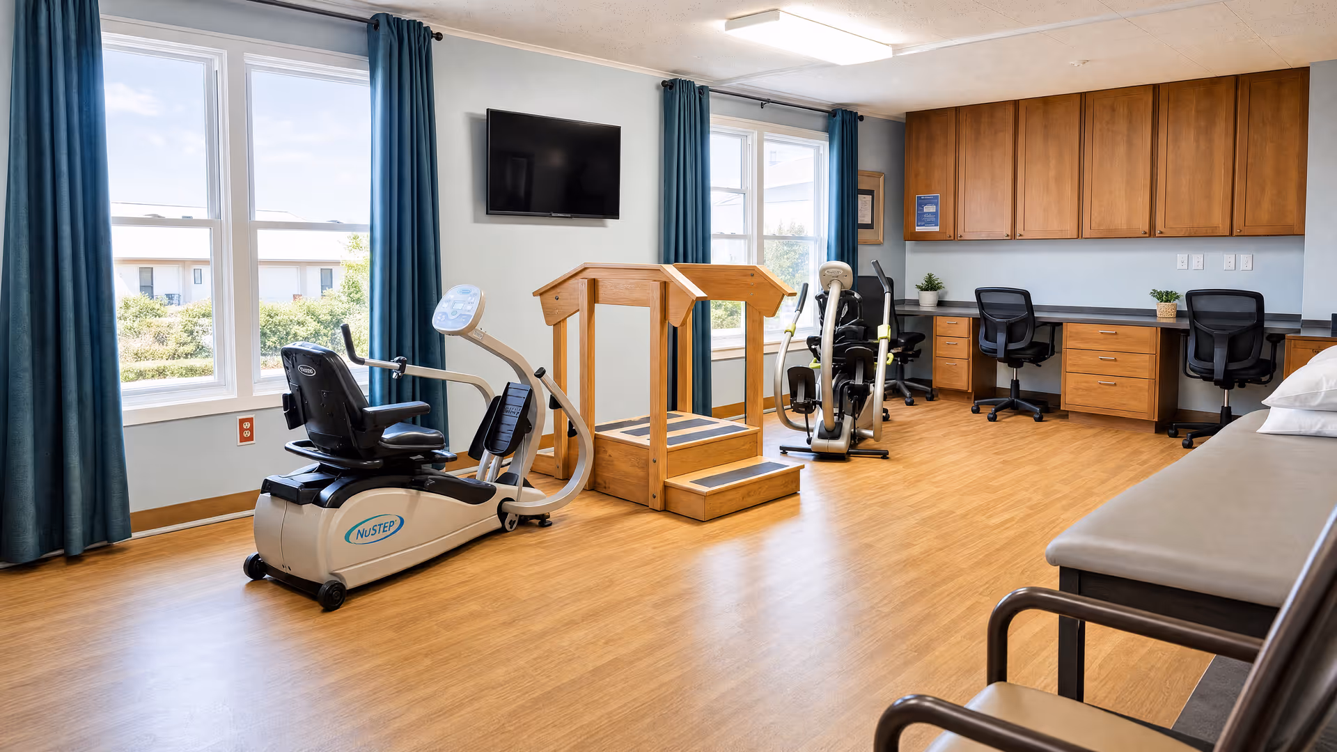 Wellness room with exercise machines, desks, cabinets, and a bed beside the large windows.