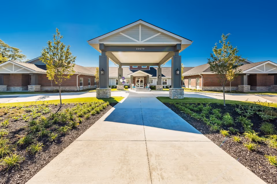 Front exterior view of Provision Living at Livonia facility showing a wide concrete walkway leading to a covered entrance with stone pillars, surrounded by landscaped garden beds and small trees under a clear blue sky.
