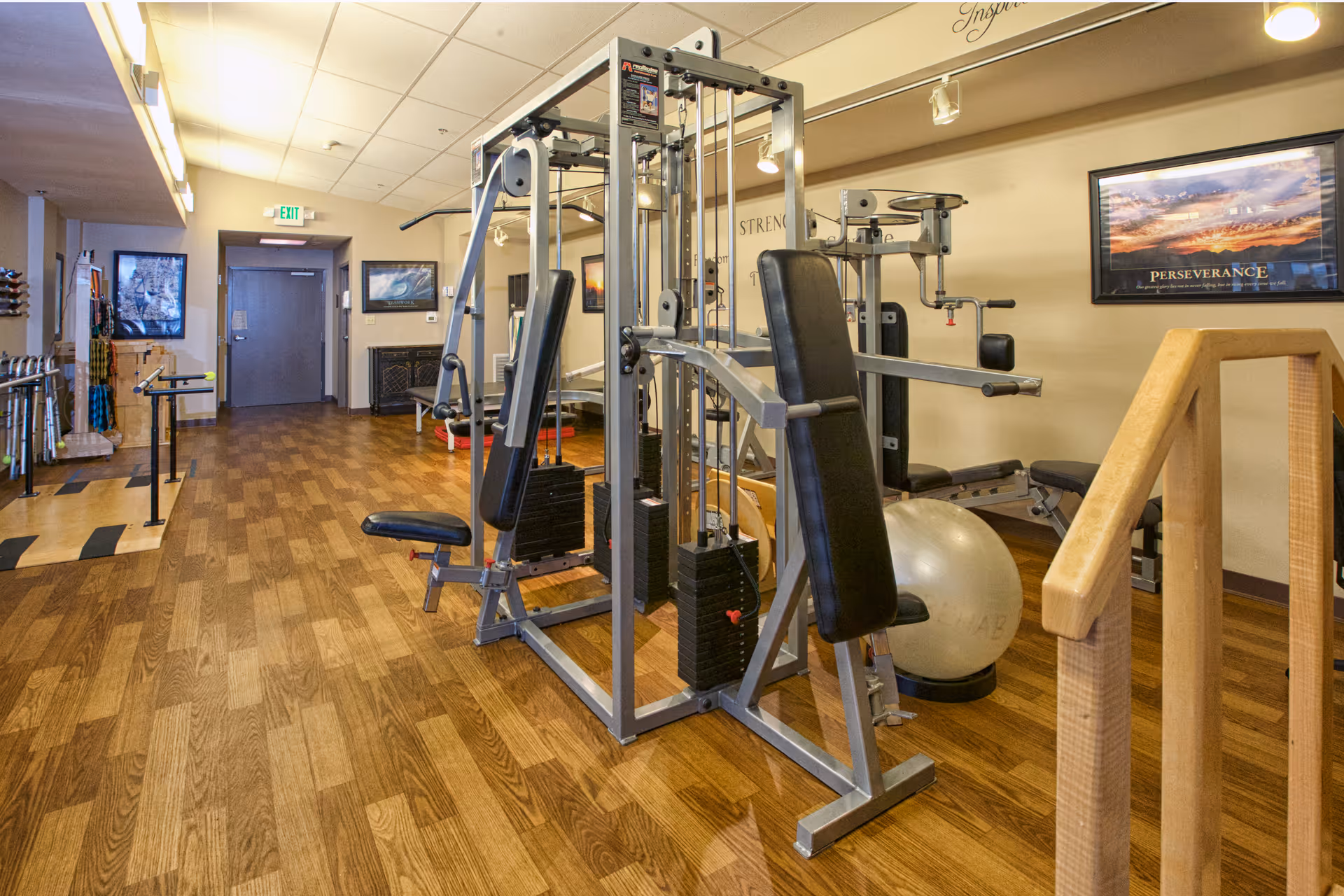Interior view of a fitness room with exercise equipment including a multi-station weight machine, exercise ball, and parallel bars. The room has wooden flooring, framed motivational posters on the walls, and an exit door at the far end.