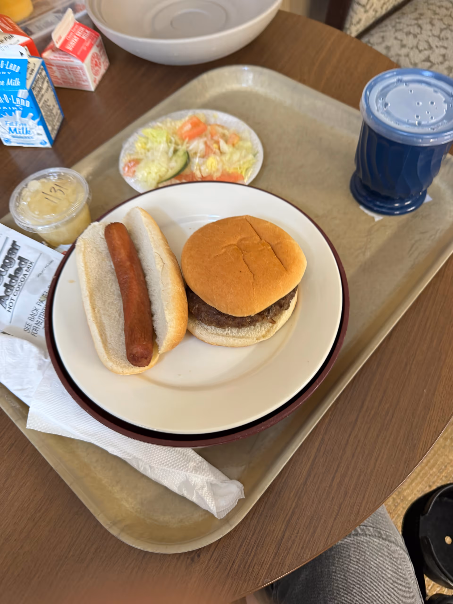 Meal tray on a table with a hamburger, hot dog, salad, and cup.