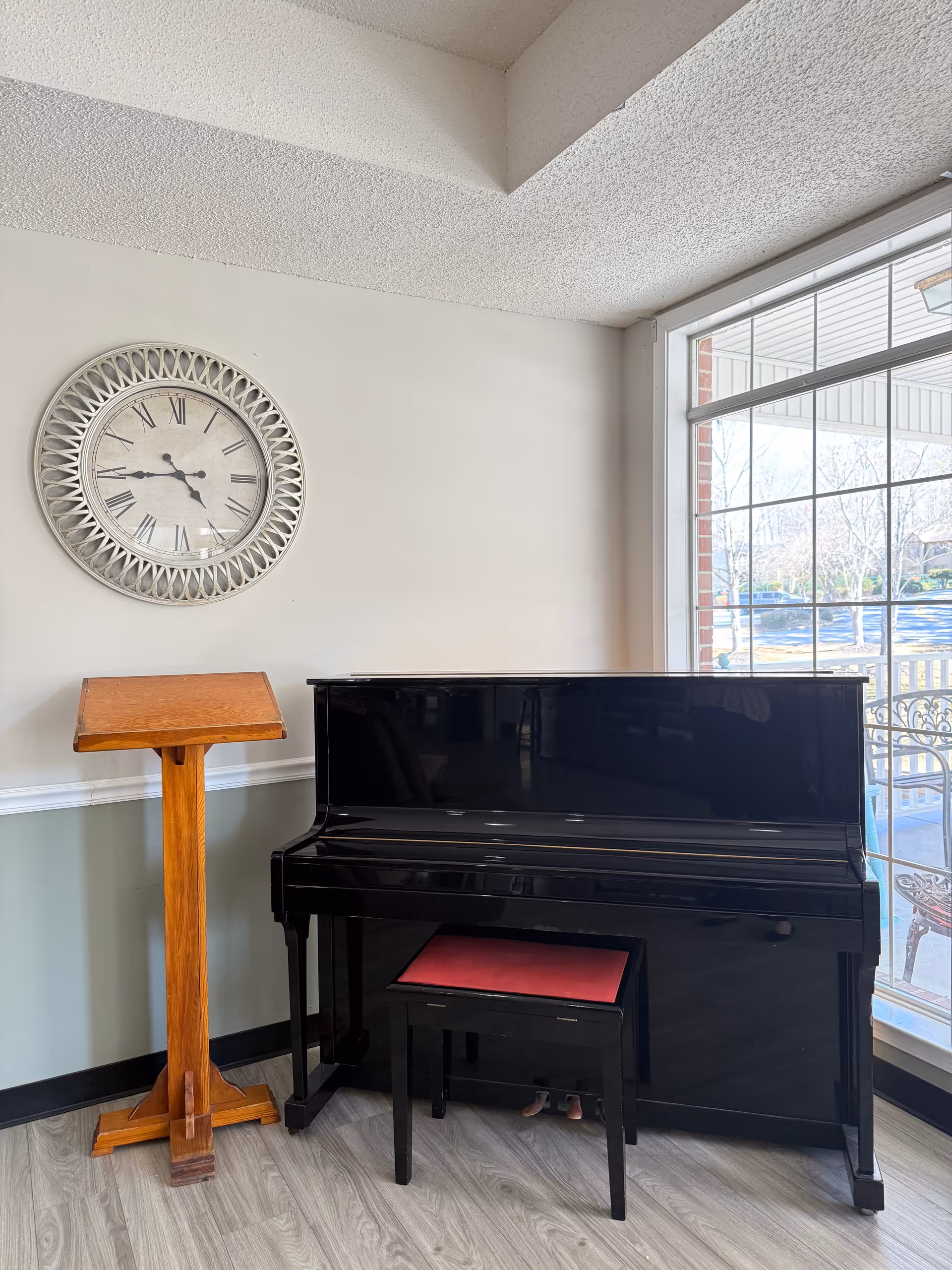 Bright living room corner with a black piano, bench, wooden lectern, wall clock, and window.