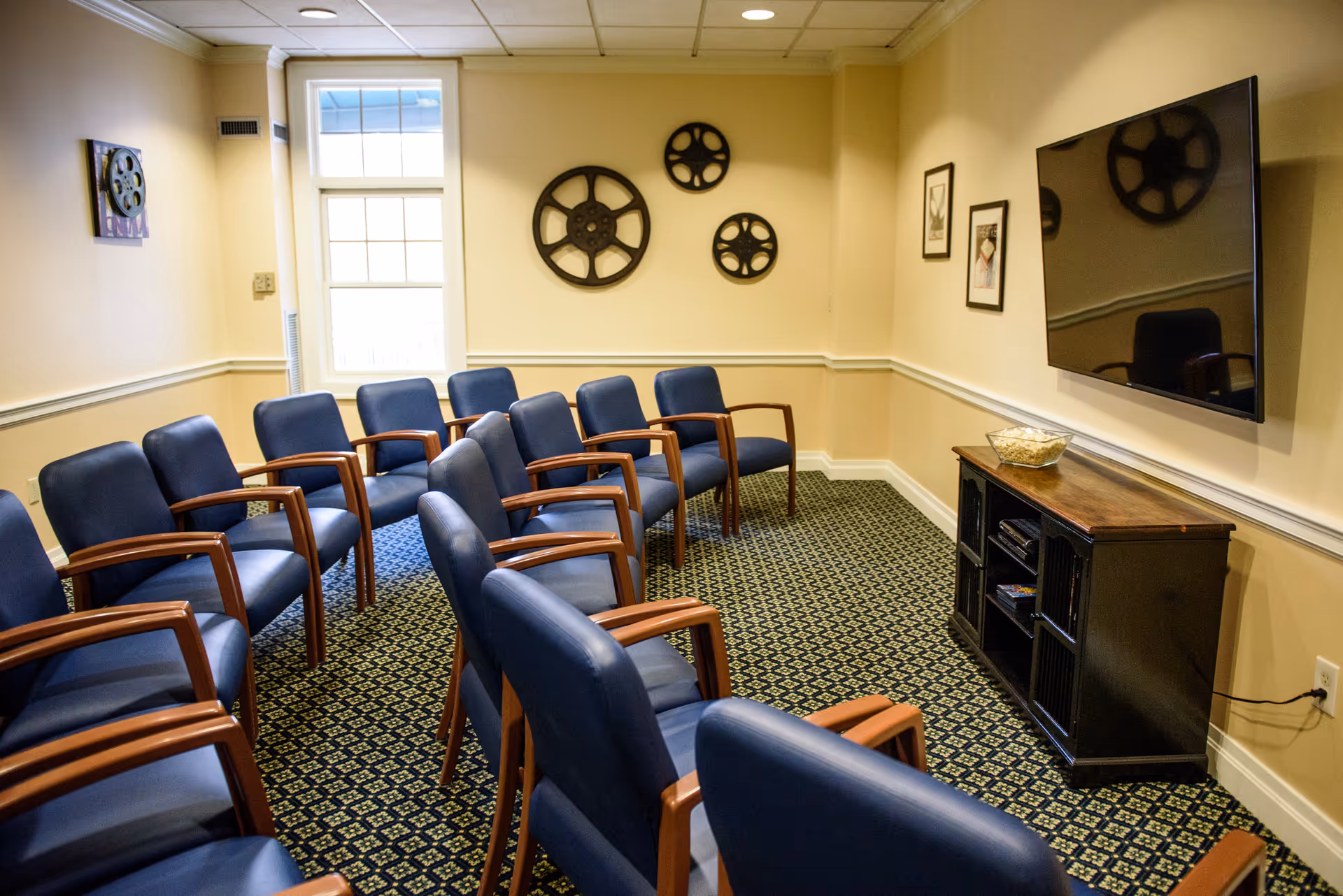 A small theater-style room with two rows of blue cushioned chairs with wooden armrests facing a large flat-screen TV mounted on a beige wall. The room has a patterned carpet, a window letting in natural light, and decorative film reels and framed pictures on the walls. A wooden cabinet below the TV holds a bowl of popcorn and some media devices.