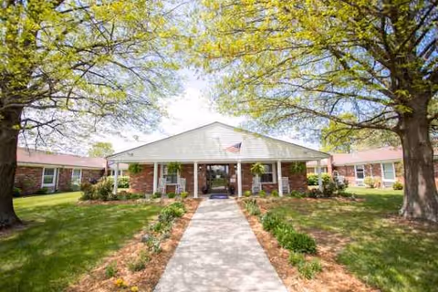 Front entrance of a single-story brick senior living facility with a walkway and trees.
