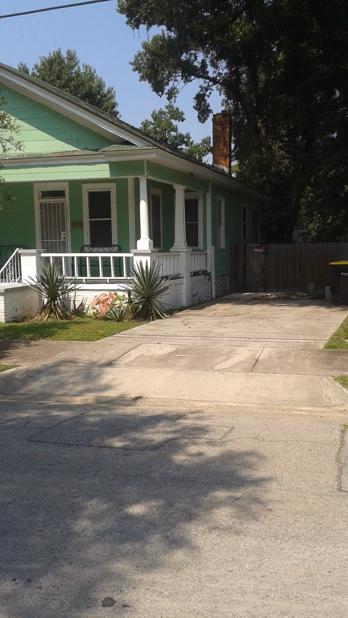 Front view of a light green single-story house with a covered porch, driveway, and small front yard plants.