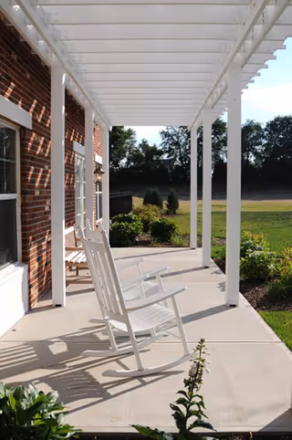 A shaded front porch with white rocking chairs under a white pergola beside a brick building overlooking a lawn.