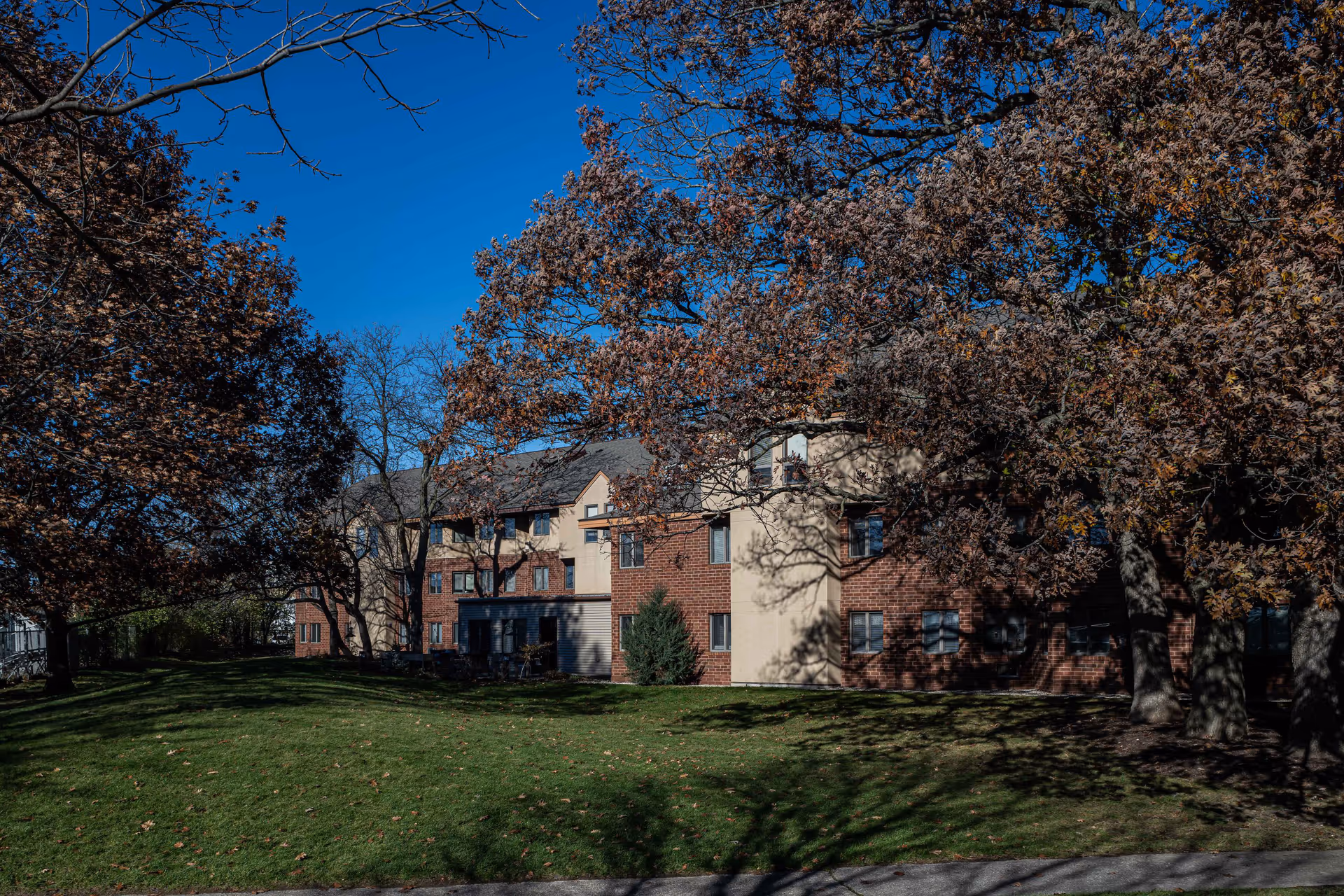 Exterior view of a multi-story senior living community building with brick and beige siding, surrounded by trees with autumn foliage and a green lawn under a clear blue sky.