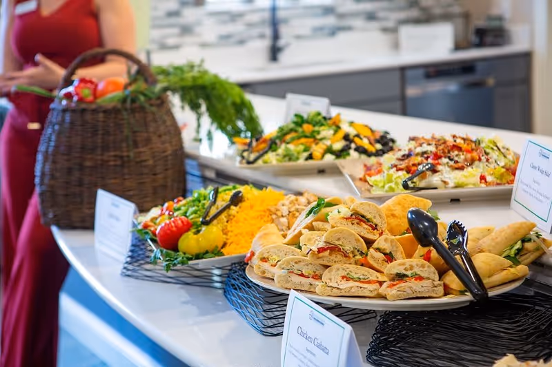 A buffet-style spread of food on a kitchen counter including sandwiches, salad, shredded cheese, and fresh vegetables with a person in a red dress in the background.