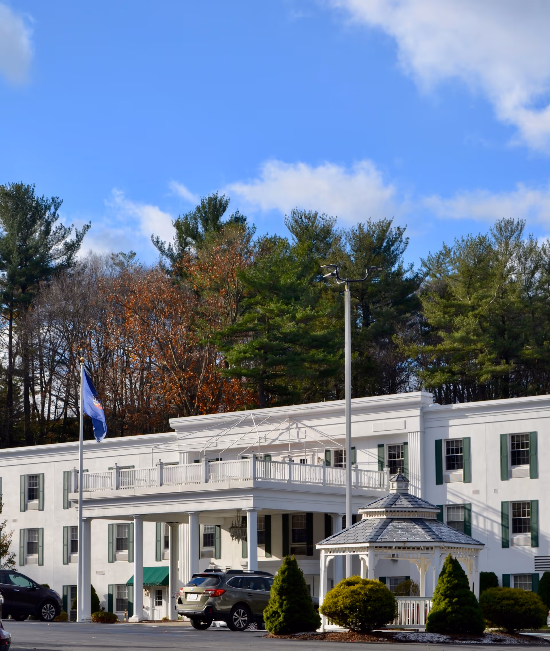 Exterior view of a white multi-story senior living facility building with green window shutters, a covered entrance supported by columns, a small white gazebo in front, several parked cars, and tall trees in the background under a blue sky with some clouds.