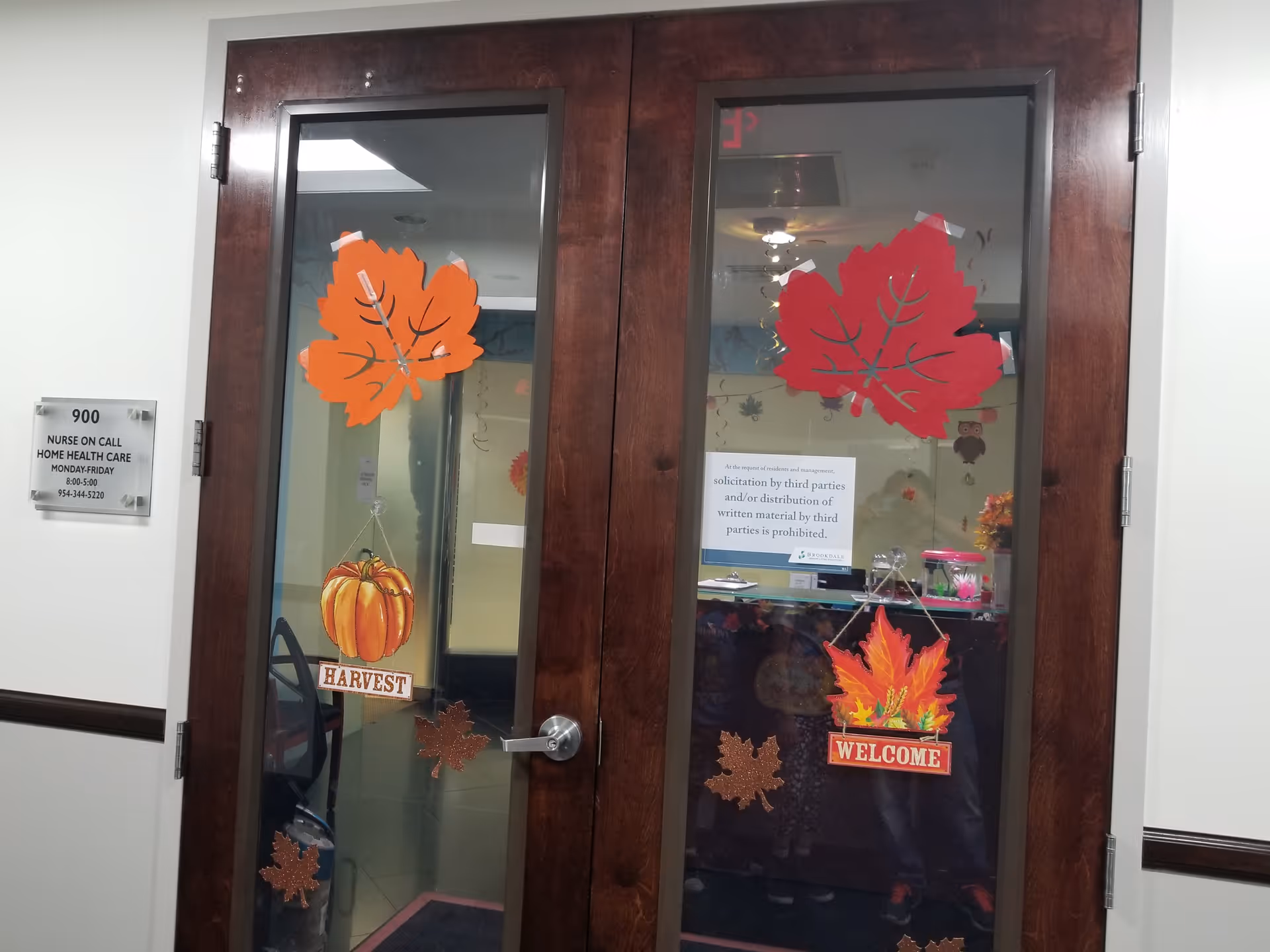 Double glass doors with autumn leaf decorations and a Harvest sign in a healthcare hallway.