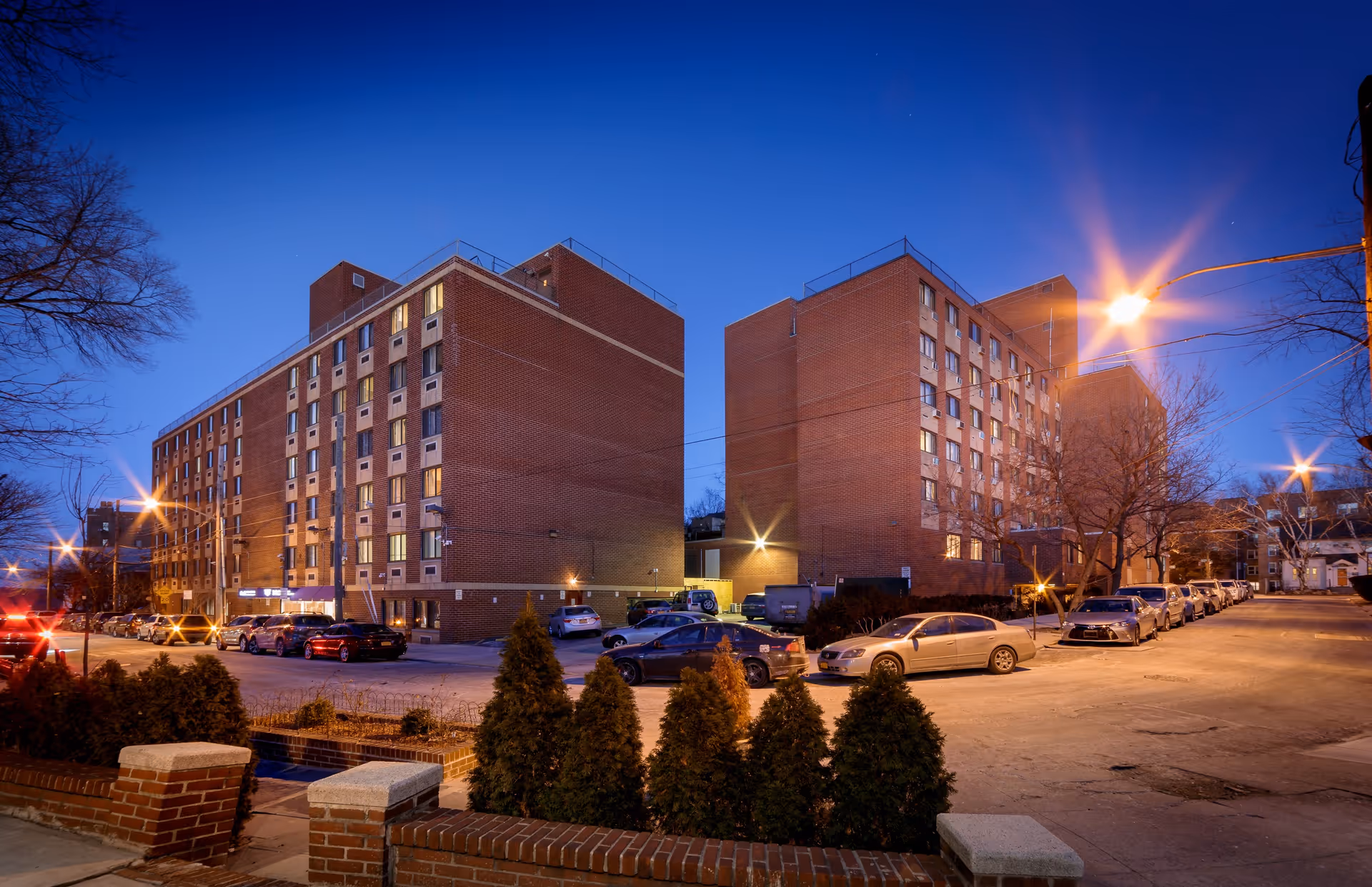 Dusk view of two connected multi-story brick buildings with a parking lot, parked cars, and illuminated streetlights.