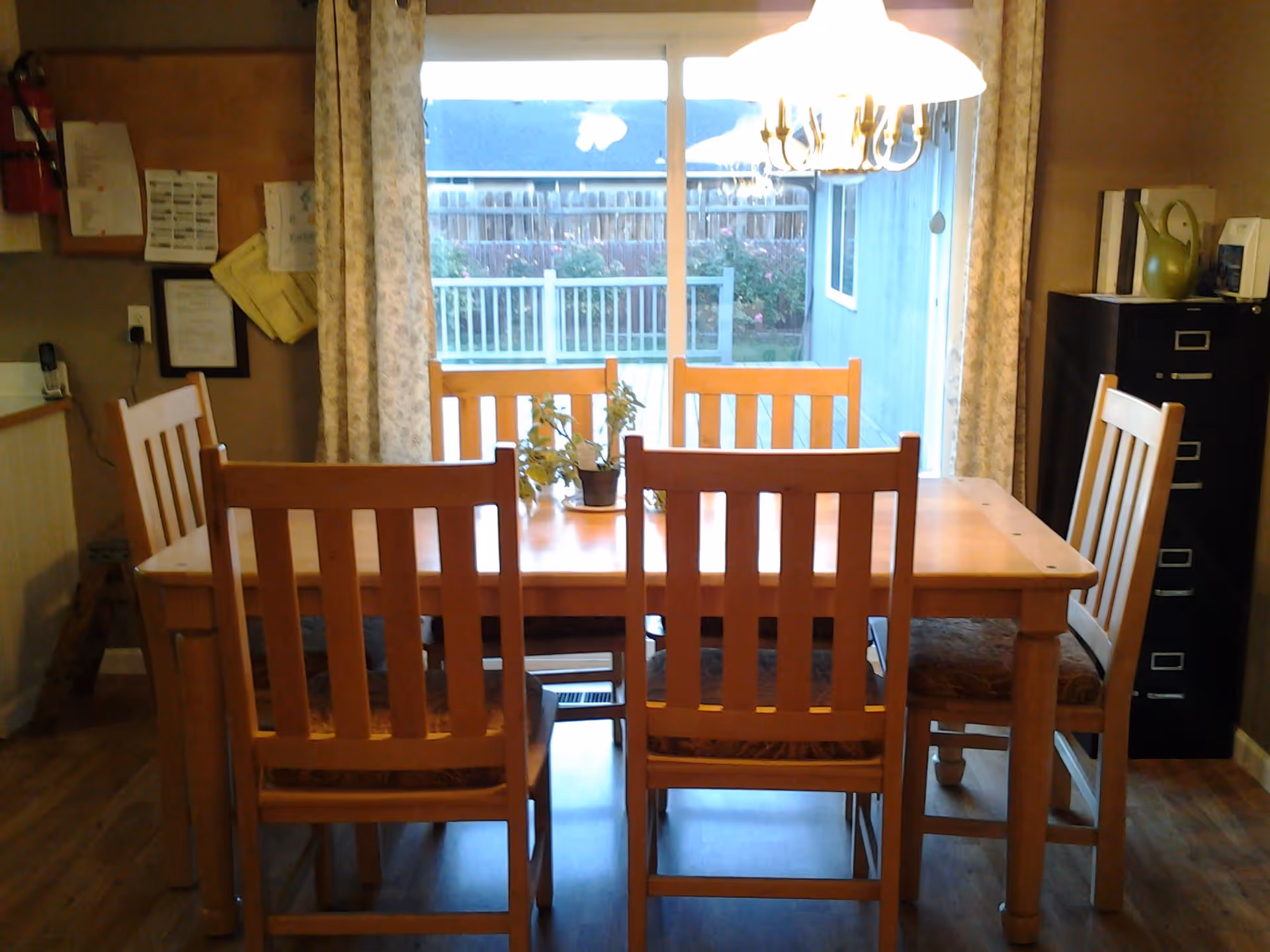 A wooden dining table with six matching wooden chairs in a room with hardwood floors. There are two small potted plants on the table. Behind the table is a large sliding glass door with curtains, showing a fenced backyard with some greenery. A chandelier light fixture hangs above the table. To the right, there is a black filing cabinet with some items on top, and to the left, a wall with various papers and a fire extinguisher mounted.