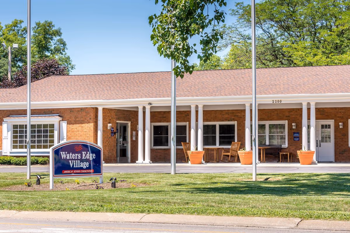 Front entrance of a single-story brick senior living building with a 'Waters Edge Village' sign, columns, potted plants, and chairs.