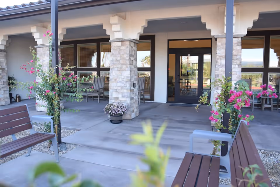 Covered front entrance of a memory care facility with stone columns, benches, potted flowers and glass doors.