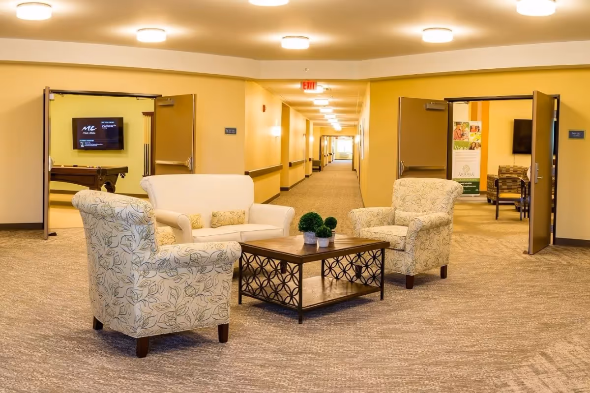 A seating area with a sofa and two patterned armchairs around a coffee table in a well-lit senior living facility corridor.
