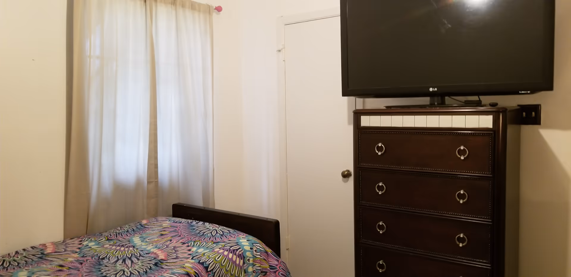 Small bedroom with bed, dresser, TV, and white curtains.