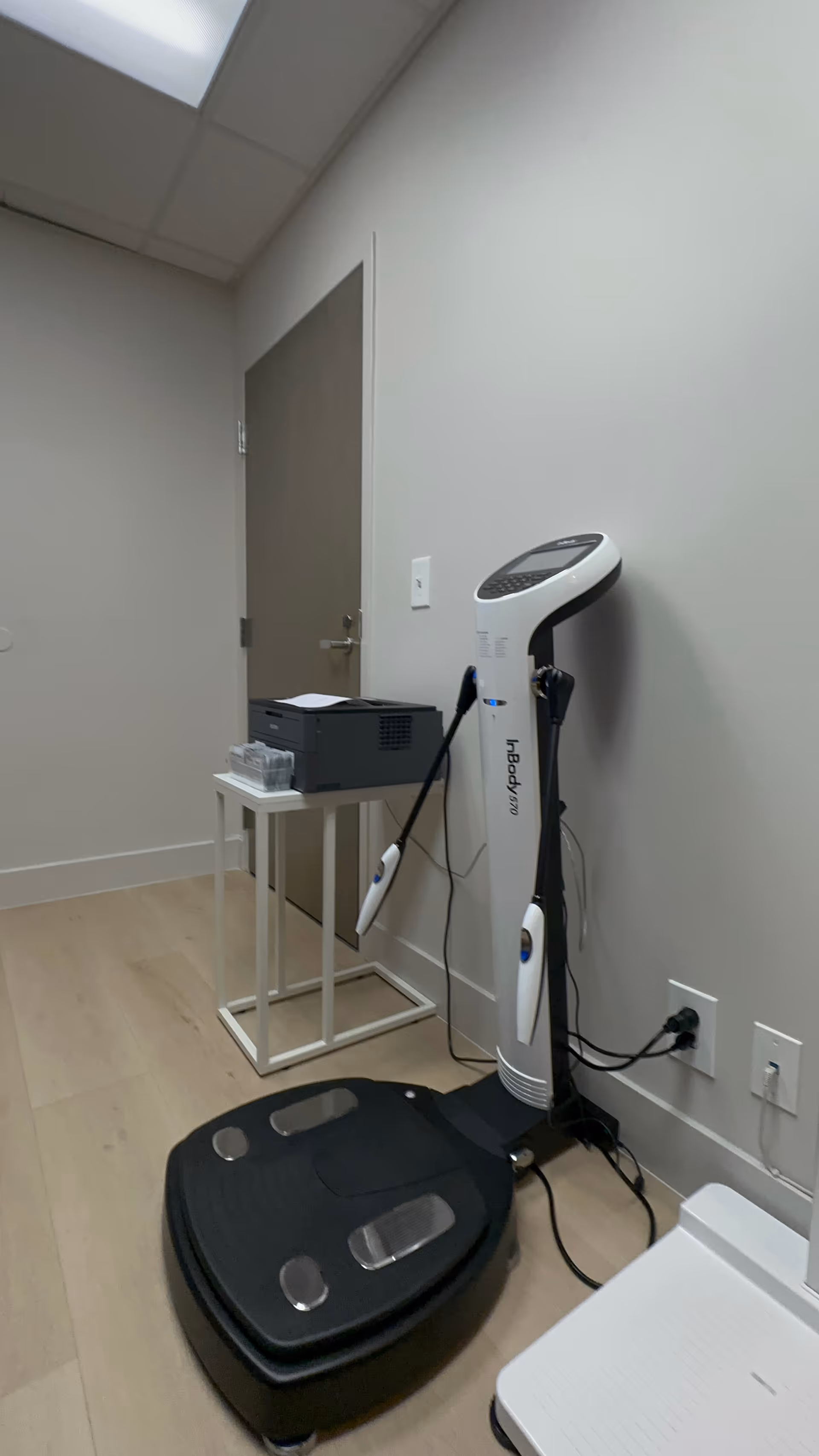 Interior clinic room with a standing body composition analyzer beside a small table and printer.