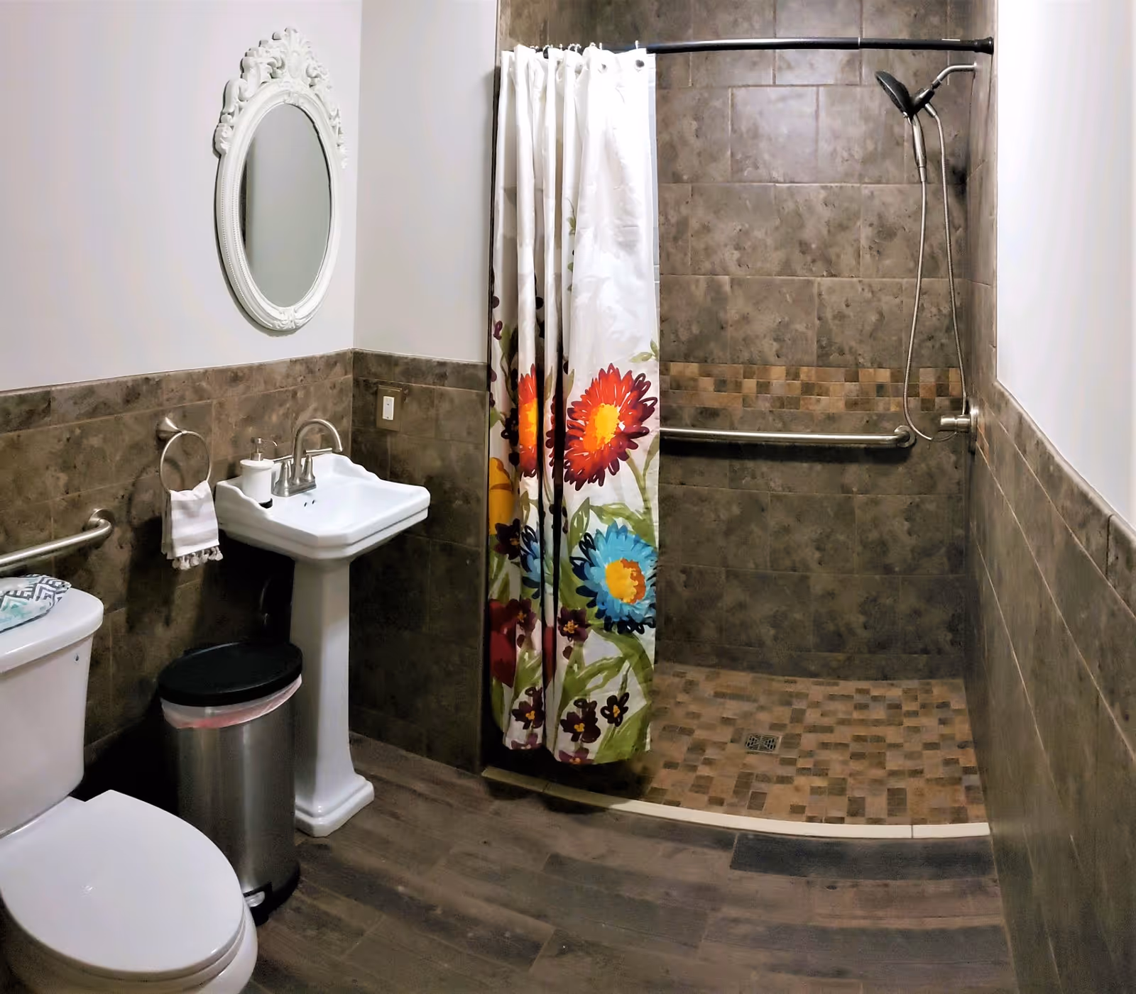 Accessible bathroom featuring a walk-in tiled shower with a colorful floral curtain, pedestal sink, toilet, and grab bars.