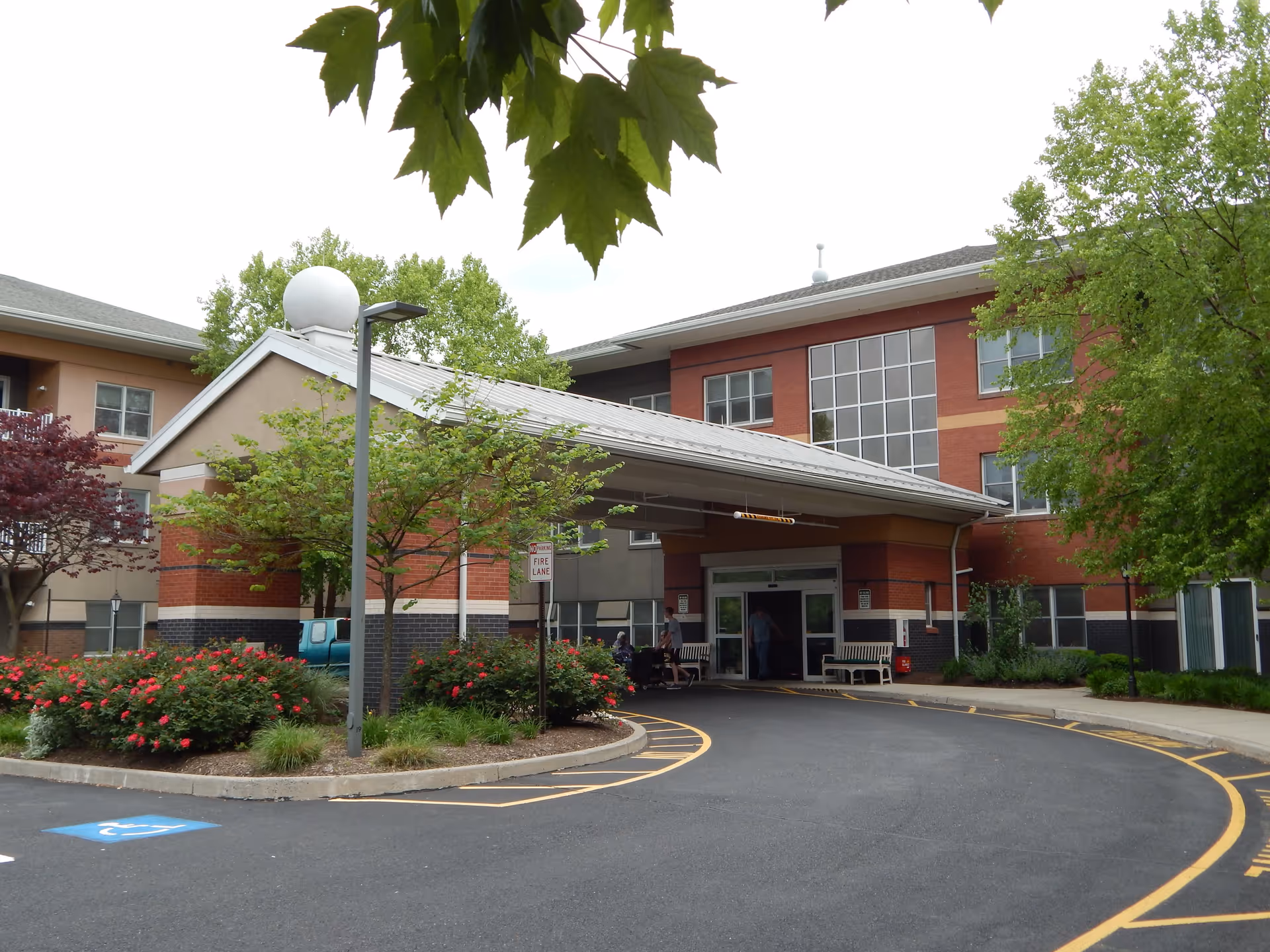 Entrance of a senior living facility with a covered drop-off area, surrounded by landscaped bushes and trees. The building has multiple floors with large windows and benches near the entrance. A few people are visible near the doorway.