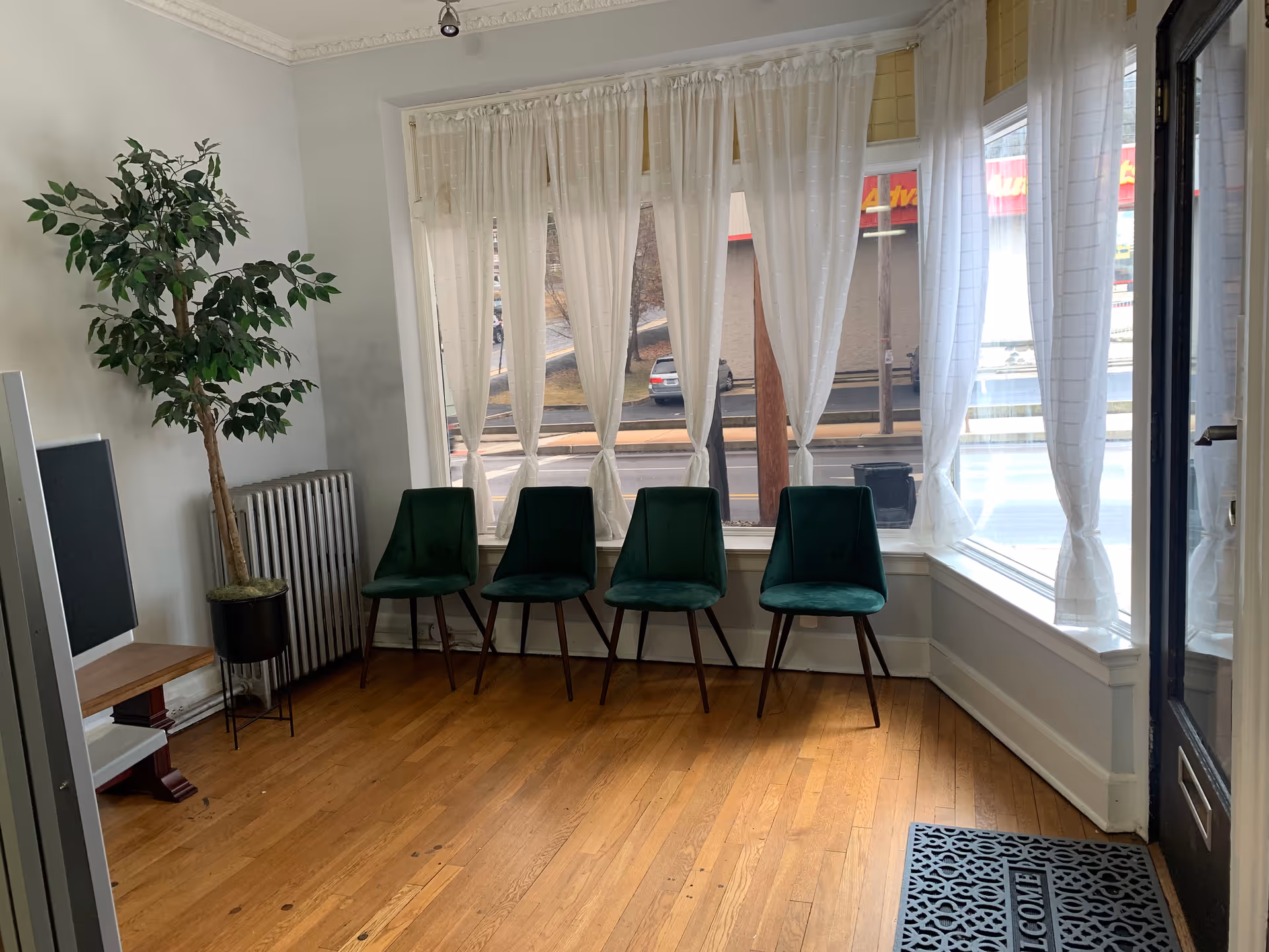 Indoor waiting area with five green chairs, a plant, and sheer-curtained windows.