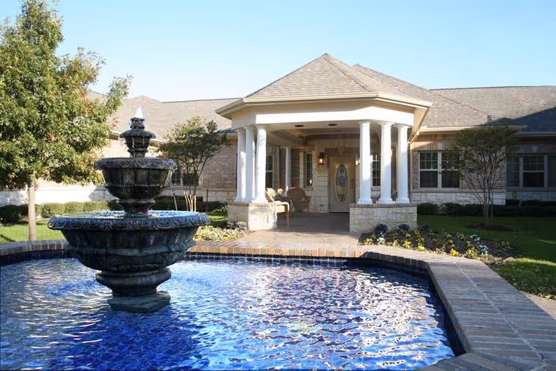 Front entrance of a single-story brick building with a covered portico and a tiered fountain in a blue-tiled pool in the foreground.
