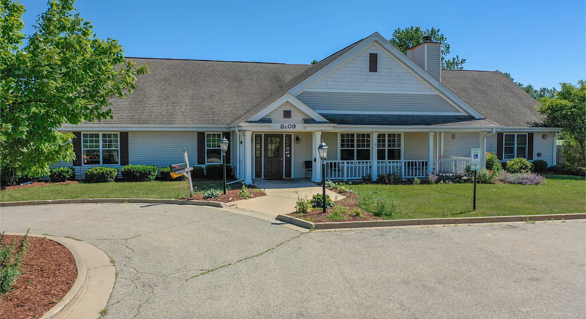 Front exterior of a single-story senior living building with a covered porch, entrance doors, lamp posts, and a landscaped driveway.