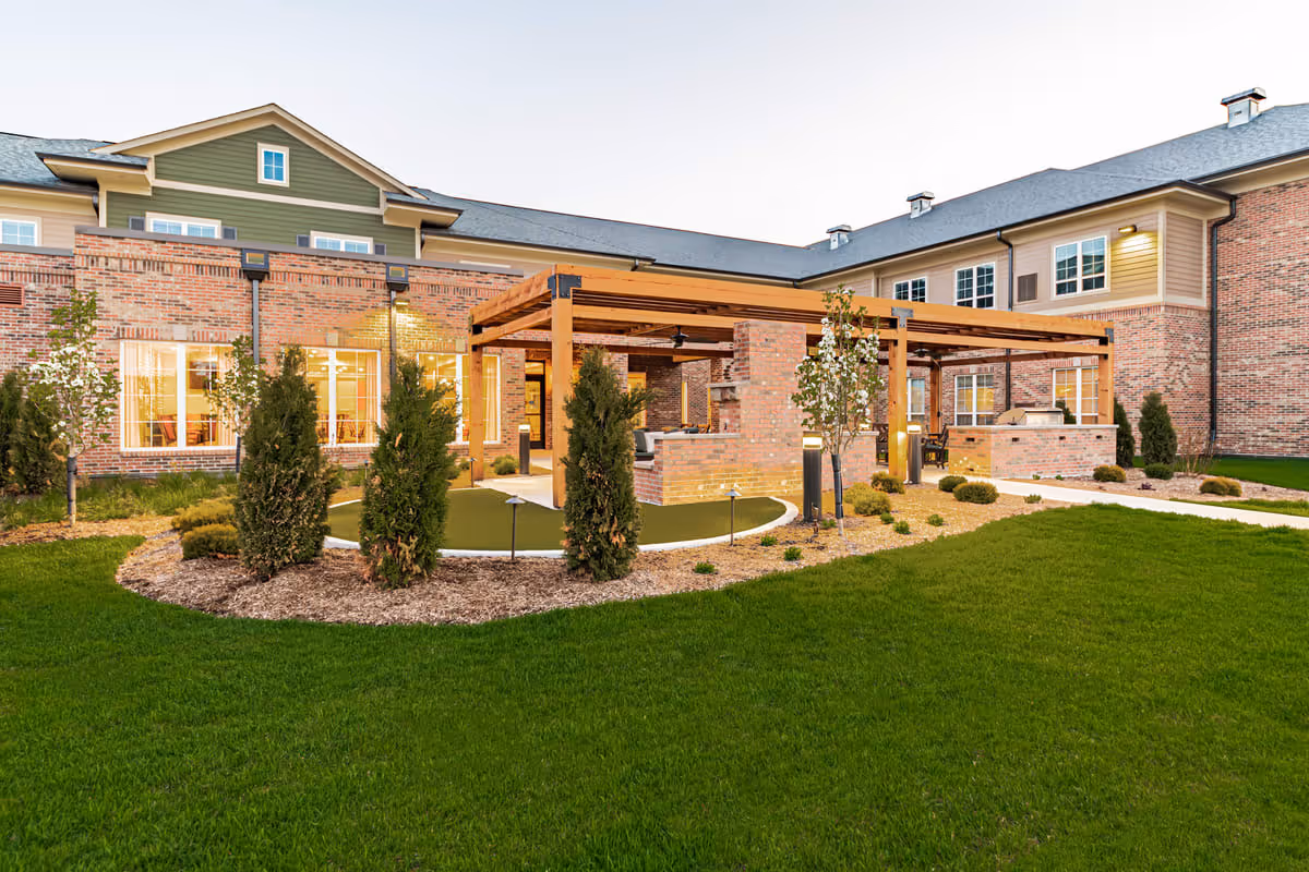 Outdoor courtyard area of a senior living facility with a green lawn, landscaped plants, and a wooden pergola structure with seating and a brick fireplace. The building exterior is made of brick with multiple windows and a green and beige upper facade.