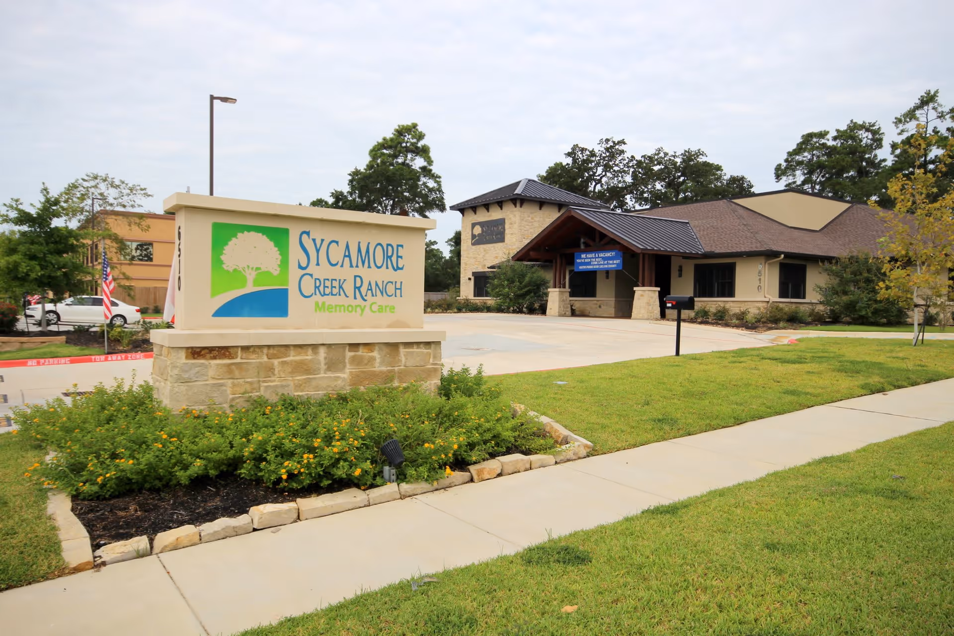 Exterior view of Sycamore Creek Ranch Memory Care facility showing the main building with stone and beige walls, a covered entrance, a parking area, and a large sign with the facility's name and logo in front surrounded by greenery and a sidewalk.