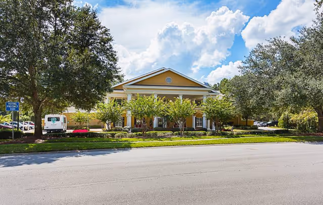 Front exterior of a yellow single-story building with columns, landscaped trees and shrubs, and a road in the foreground.