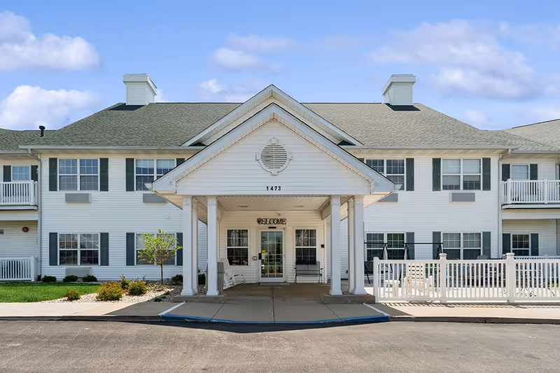 Front exterior view of McKay Manor, a two-story white building with green shutters and a covered entrance with a 'WELCOME' sign above the door. There are small trees and shrubs near the entrance and a paved driveway in front.