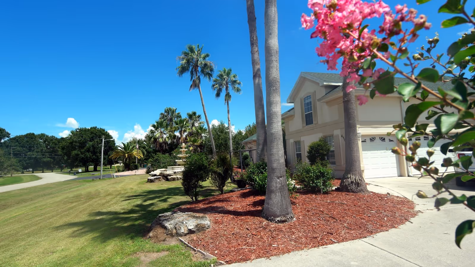 Front exterior of a two-story house with palm trees, landscaped mulch beds, a driveway and garage under a bright blue sky.