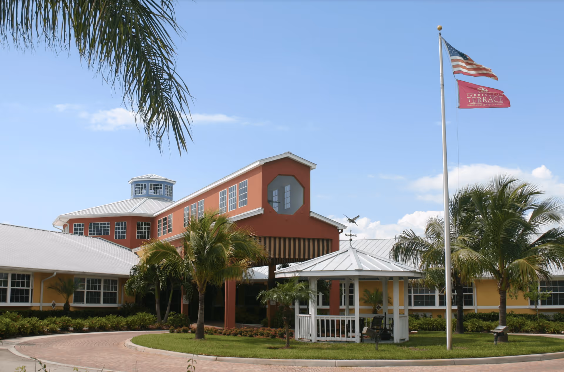 Exterior view of a senior living facility with a circular driveway, palm trees, a small white gazebo, and two flags on a flagpole against a clear blue sky.