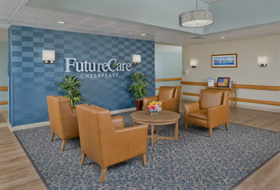Reception lounge with four tan leather armchairs around a round wooden table in front of a blue tiled wall reading 'FutureCare Chesapeake'.