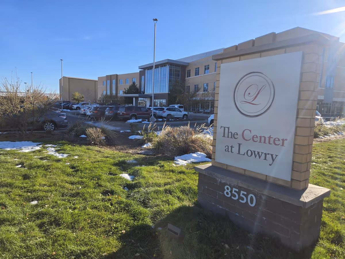 Entrance sign reading "The Center at Lowry" with the address 8550 in front of a three-story care facility, parking lot, and grassy foreground with patches of snow.
