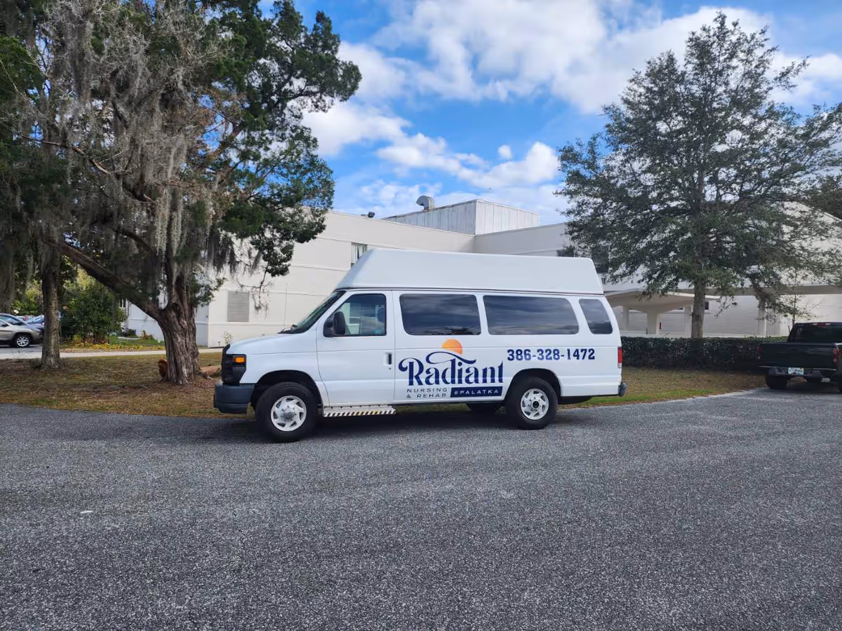 A white transport van parked in a parking lot next to a building. The van has the logo and contact number of Radiant Nursing and Rehab at Palatka on its side. There are trees and a partly cloudy sky in the background.