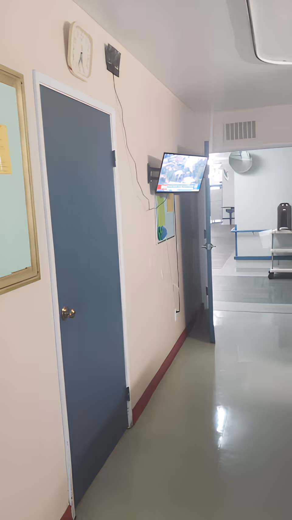 Interior hallway of a senior living facility with a closed blue door, wall clock, mounted TV, and an open doorway leading to a common area.