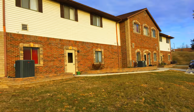 Two-story brick-and-siding building front with multiple windows and doors, outdoor HVAC units, a walkway, and a grassy lawn under a blue sky.