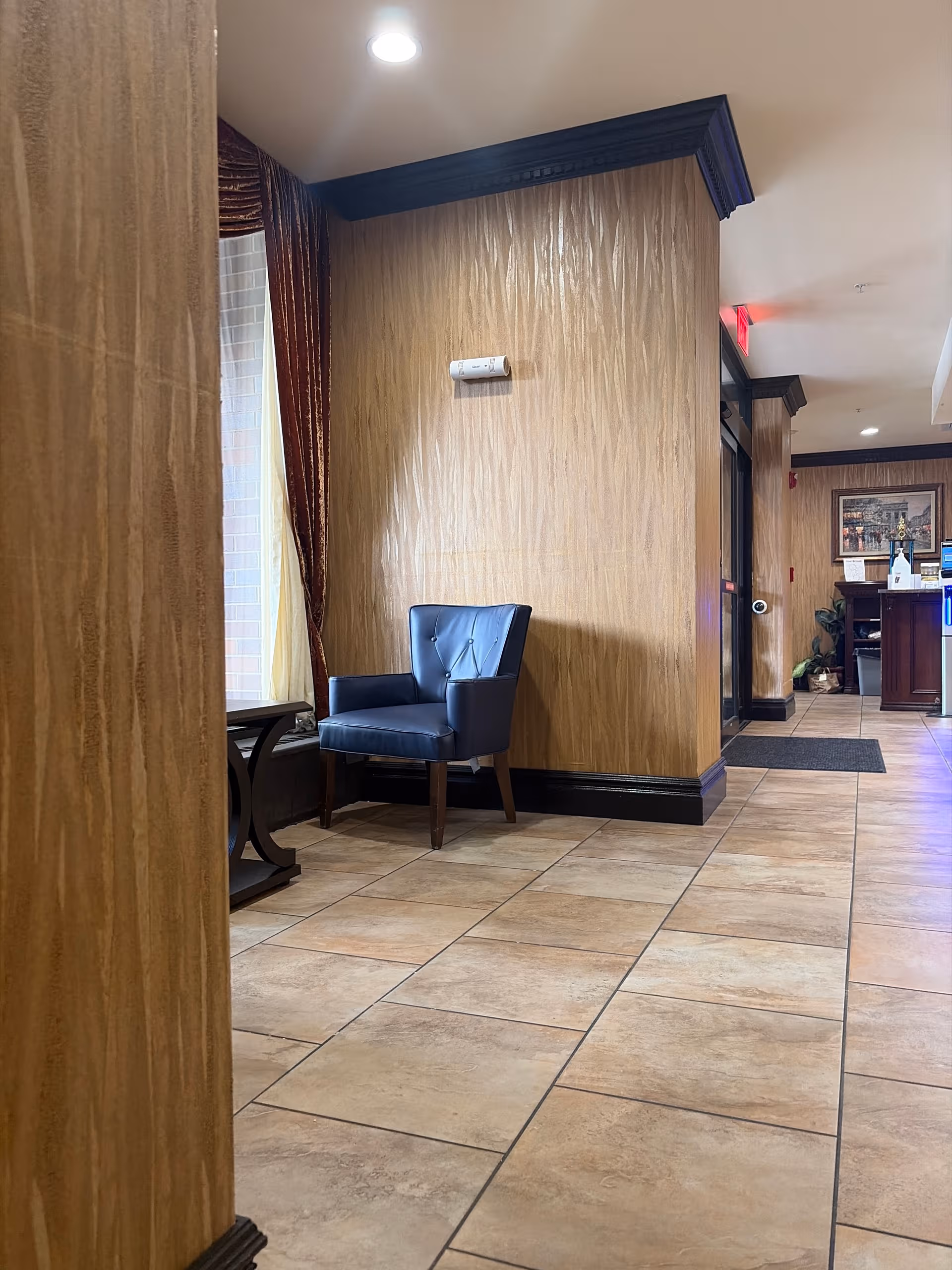 Interior lobby area with a blue chair, tiled floor, wood-paneled walls, and a hallway.
