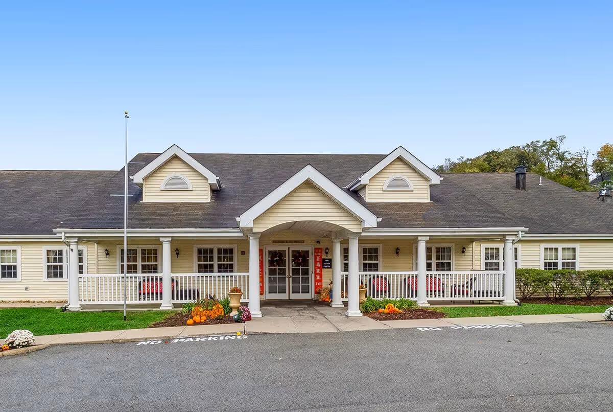 Front exterior view of a single-story senior living facility building with a covered entrance supported by white columns. The building has beige siding, a dark shingled roof with two dormer windows, and a porch with white railings. There are pumpkins and fall decorations near the entrance, and a 'No Parking' sign painted on the driveway.