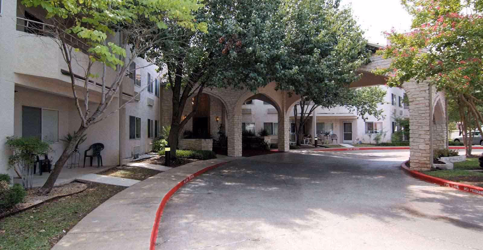 Exterior view of Solstice Senior Living at Austin showing a driveway with a covered entrance supported by stone arches, surrounded by trees and shrubs, with the building visible in the background.