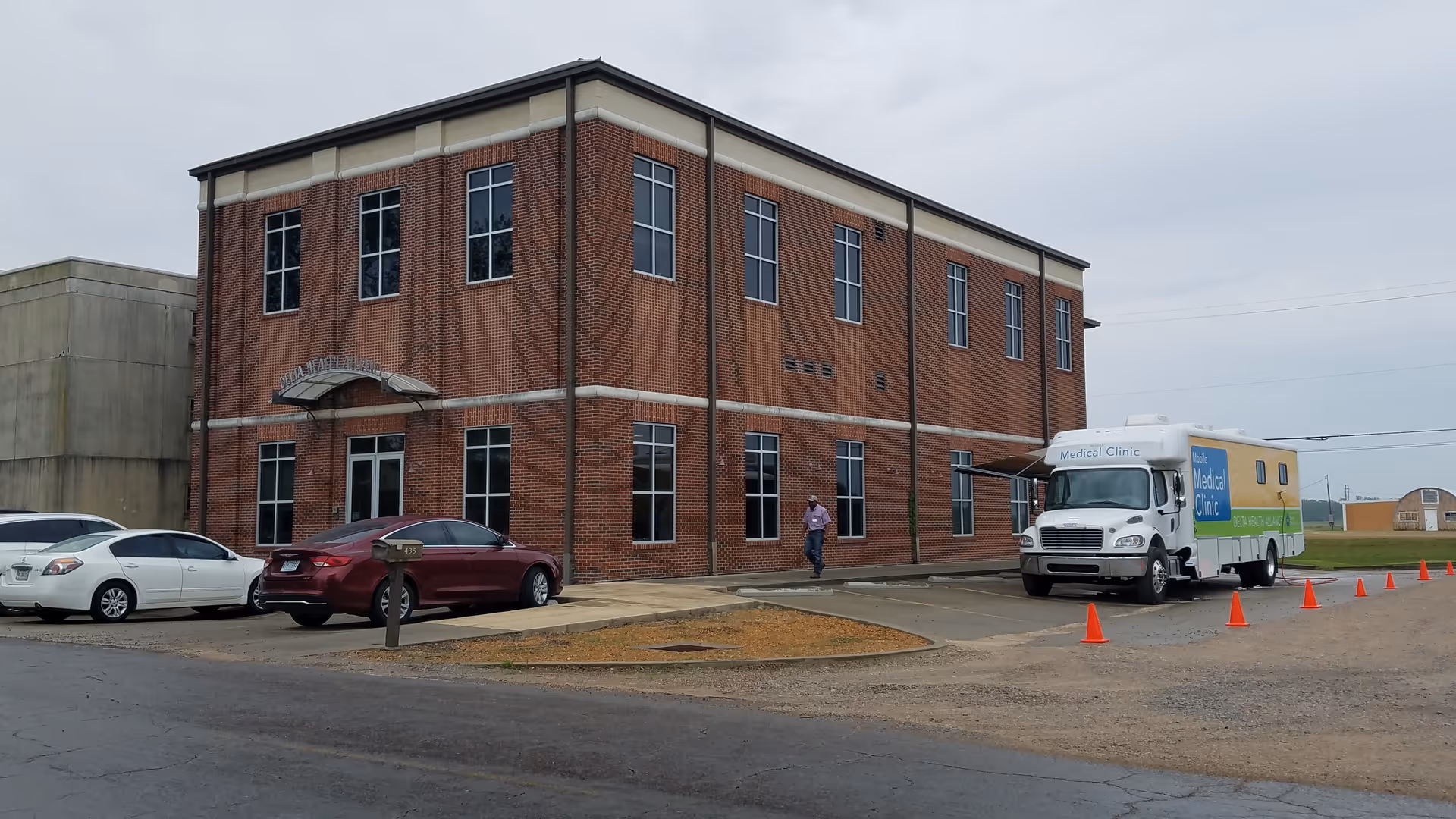 Brick two-story Delta Health Alliance facility exterior with parked cars and an ambulance nearby.