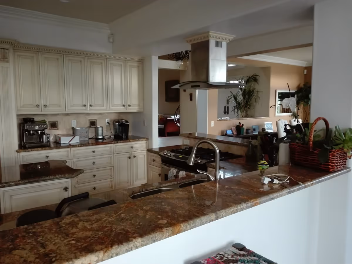 Open kitchen with marble countertops, cream cabinets, a stainless steel range hood and sink, looking into an adjacent living area.