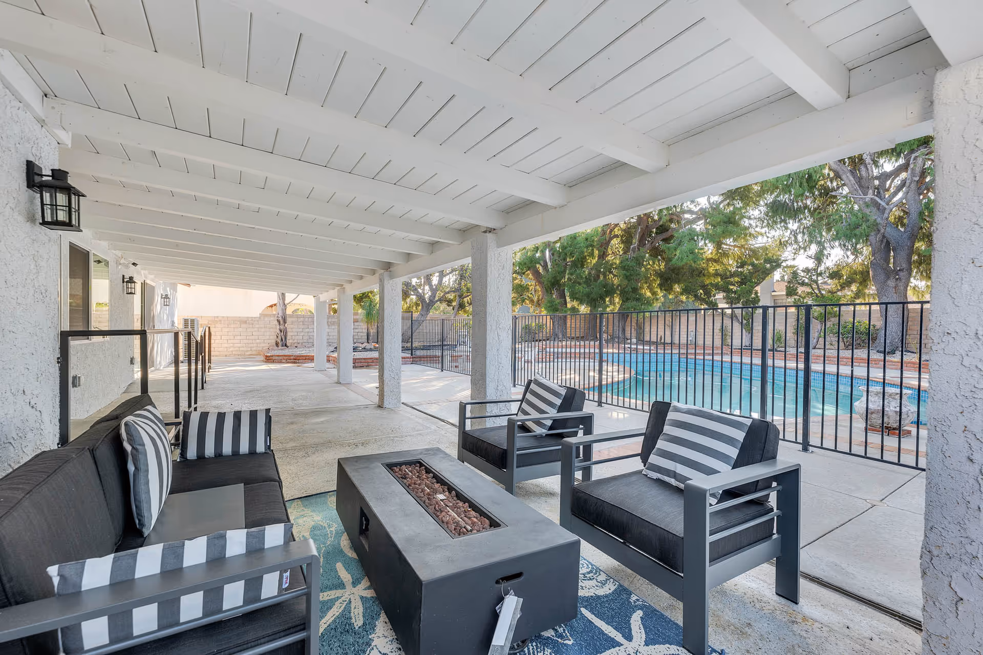 Covered outdoor seating area with modern lounge chairs and a pool beyond a fence.