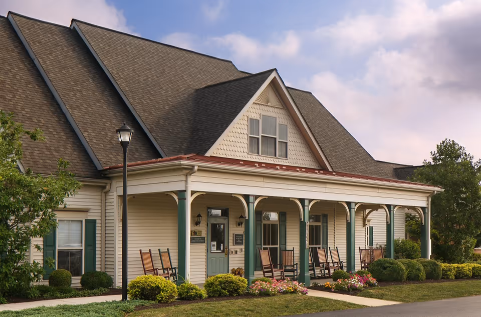 Front exterior of a cottage-style senior living building with a covered porch, rocking chairs, and landscaped shrubs and flowers.