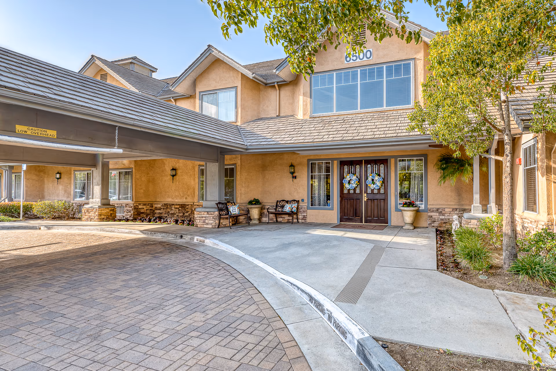Front entrance and covered porte-cochere of a senior living building with double decorative doors, benches, and potted plants.