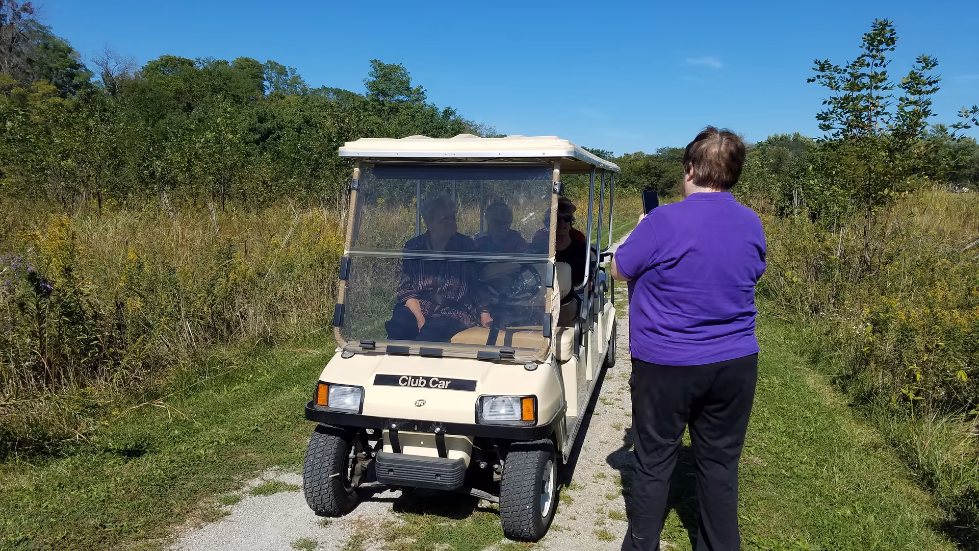 Residents ride in a golf cart on an outdoor trail while a staff member photographs them.
