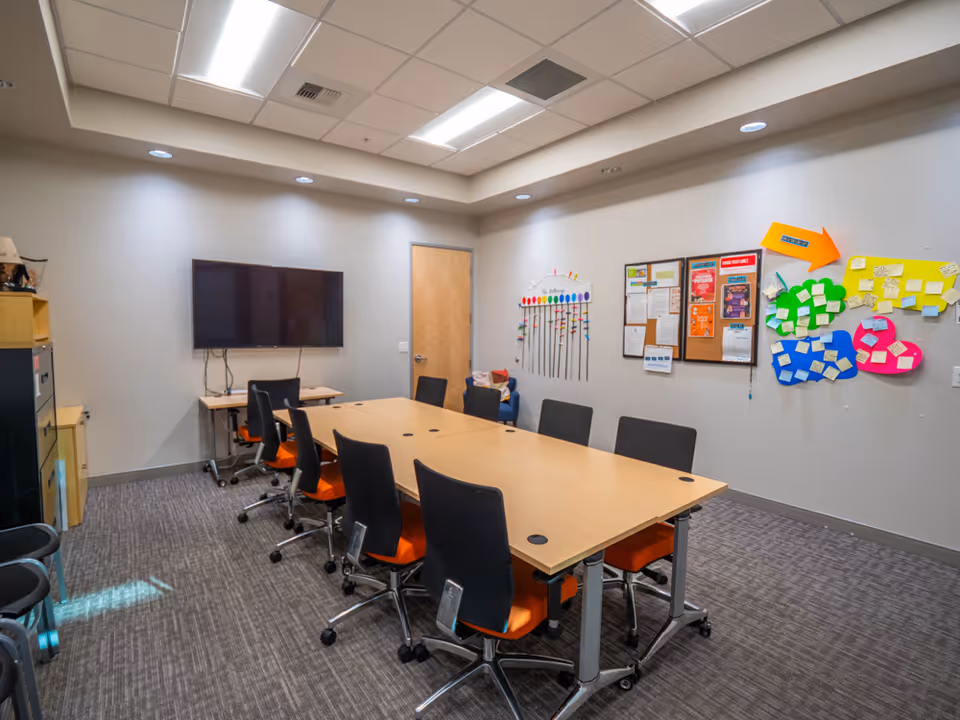 A conference-style classroom with a long table, chairs, a wall-mounted TV, and bulletin boards.