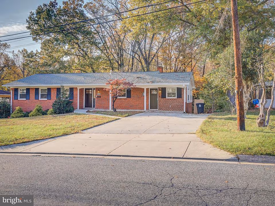 Front exterior of a single-story brick house with a driveway, lawn, and trees in the background.